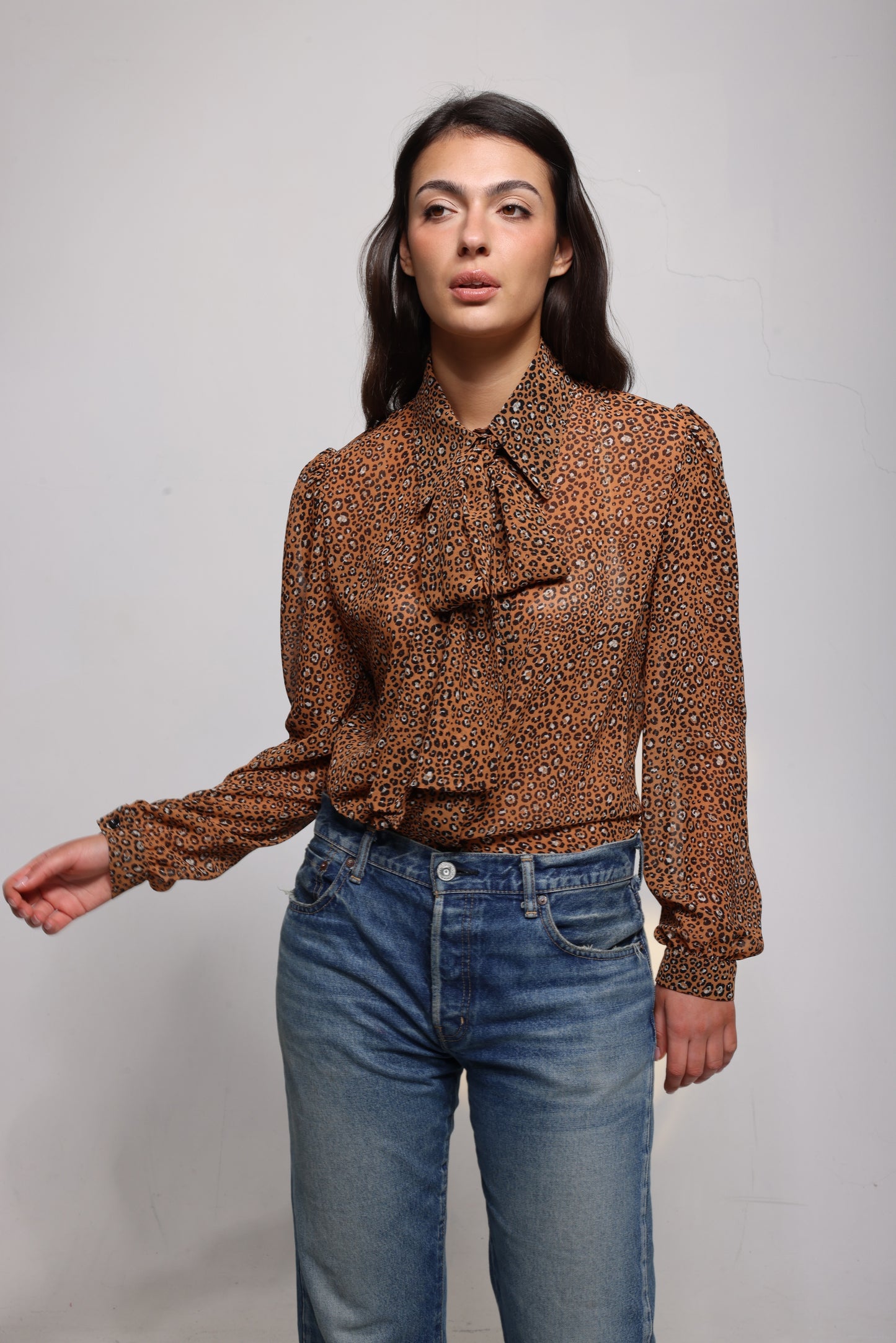 A woman modeling a brown and black leopard-print long-sleeve blouse with a ruffled tie-neck collar, paired with blue jeans, against a plain light gray studio background.
