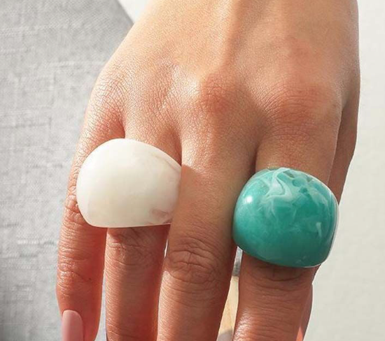 Hand wearing two rings, one white and one turquoise, on a neutral background
