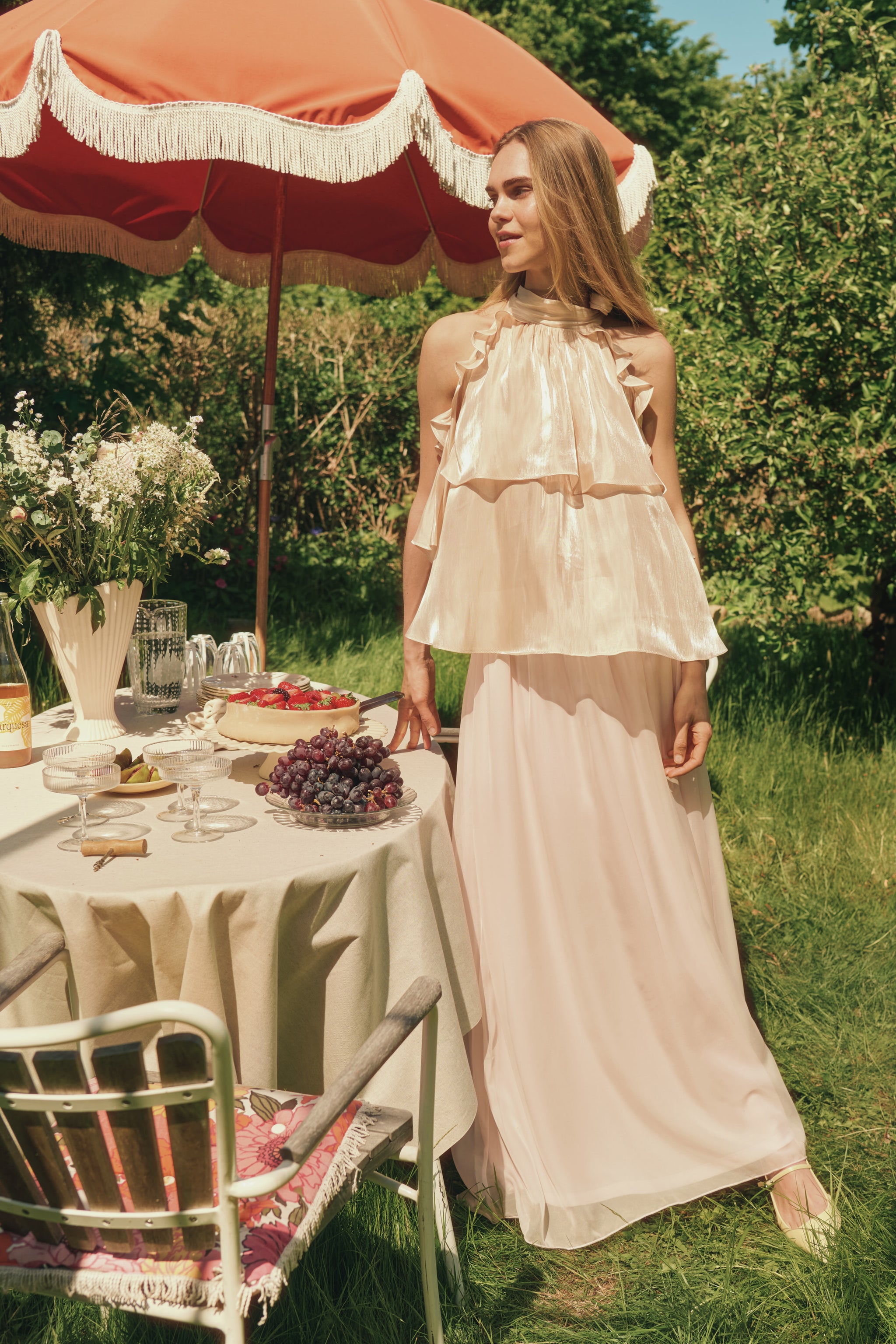 Woman in a white dress standing outdoors near a table with food and a pink umbrella.