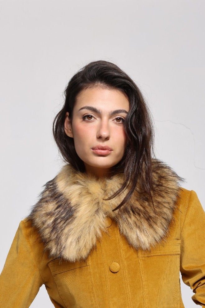 A woman modeling a mustard yellow corduroy jacket and matching skirt set, featuring a large brown and beige faux fur collar on the jacket, against a plain light gray background.