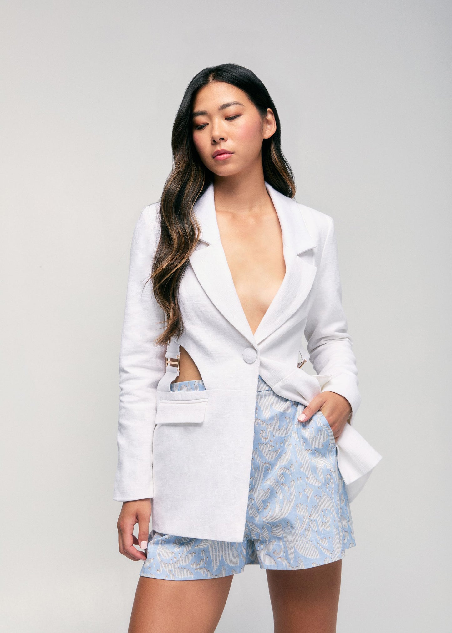 Woman wearing a white blazer and light blue floral shorts on a gray background