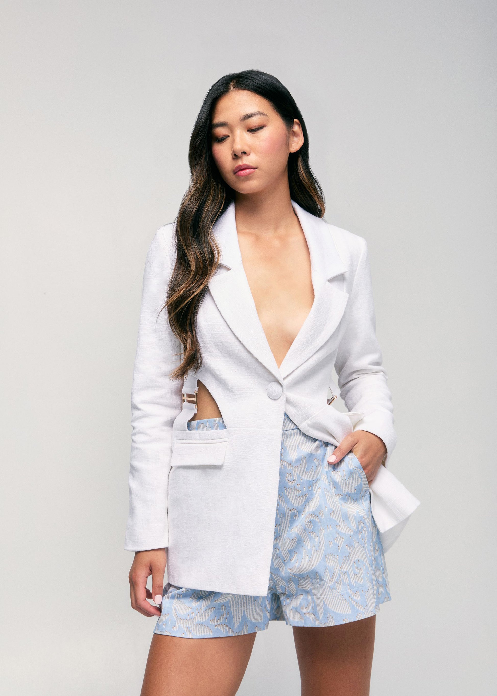 Woman wearing a white blazer and light blue floral shorts on a gray background