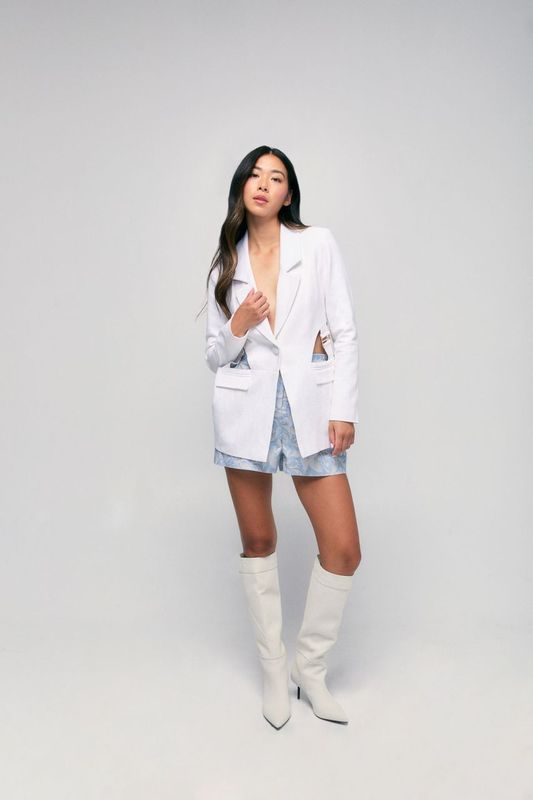 Woman wearing a white blazer, light blue dress, and white knee-high boots on a plain background
